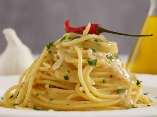 Spaghetti conditi con aglio, olio e peperoncino, guarniti con un peperoncino rosso e prezzemolo. Sullo sfondo, un bulbo d'aglio e una bottiglia d'olio.