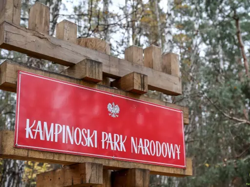 Drewniany drogowskaz z napisem "KAMPINOSKI PARK NARODOWY" i godłem Polski. W tle las.