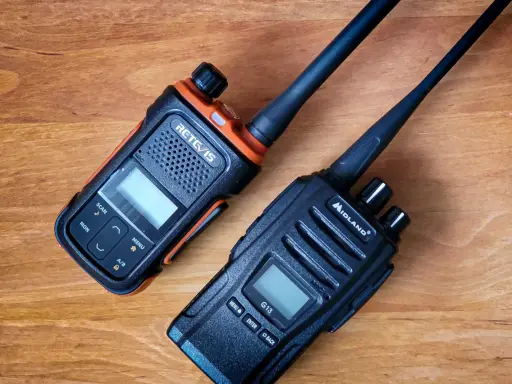 Dwa radiotelefony, jeden pomarańczowo-czarny RETCIS, drugi czarny Midland, leżą na drewnianym blacie.