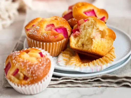 Saftige Rhabarber Muffins, mit Puderzucker bestäubt, liegen auf einem Teller. Einer ist angeschnitten und zeigt die rosa Rhabarberstücke.