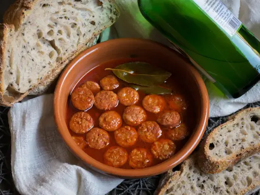 Chorizo a la sidra en cazuela de barro, acompañado de pan rústico y una botella de sidra verde.