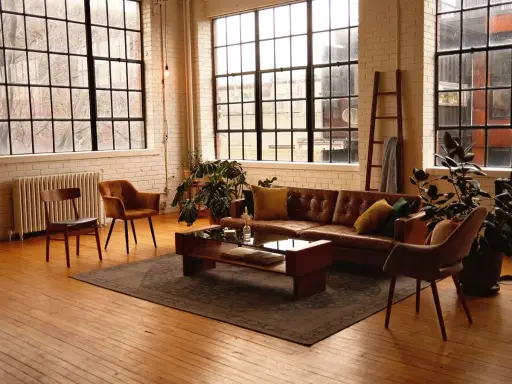Przytulny salon w stylu loft aranżacje. Brązowa skórzana kanapa, drewniane meble i duże okna tworzą ciepłą atmosferę.