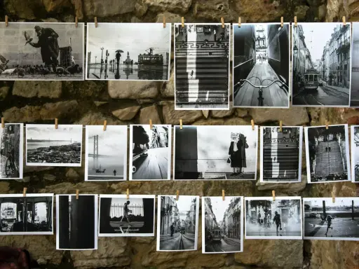 Czarno-białe fotografie wiszą na sznurku na kamiennej ścianie. Widać na nich uliczne sceny z Lizbony, w tym tramwaje, most i ludzi.