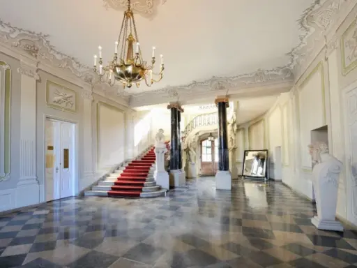 Elegancka klatka schodowa z czerwonym dywanem i żyrandolem w Pałacu Branickich w Białymstoku. Sprawdź cennik zwiedzania!