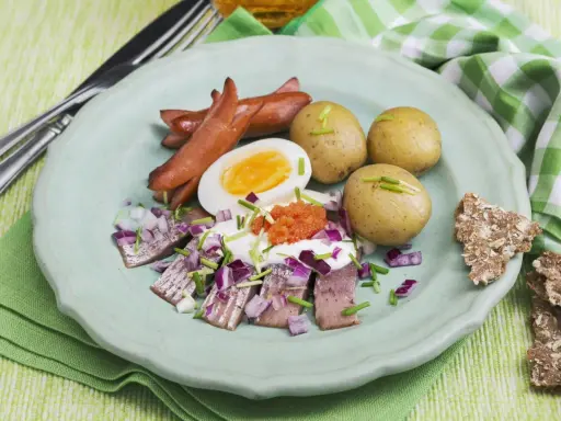 En tallrik med sill, ägg, prinskorv, potatis och rom. En härlig påskbord mat som ser utsökt ut.