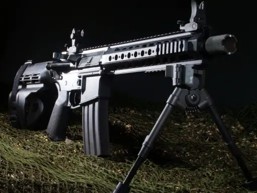 Czarny karabin AR z magazynkiem bębnowym i dwójnogiem, leżący na zielonej siatce maskującej.