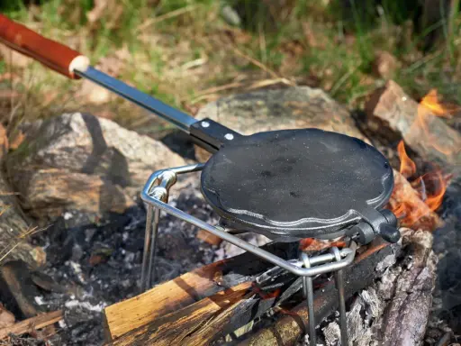En gjutjärnspanna med trähandtag står på en ställning över en öppen eld. Perfekt för att steka plättar eller våfflor ute i naturen.
