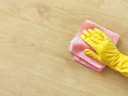 En hand i gul handske torkar av ett ljust trägolv med en rosa trasa. Perfekt för pigmenterad olja till ekbord.