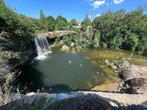 Gente disfrutando de un día soleado en las pozas del río Tus, con una cascada y vegetación exuberante.