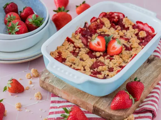 Ein frisch gebackener Erdbeer Crumble in einer hellblauen Auflaufform, garniert mit Erdbeerhälften. Daneben eine Schüssel voller Erdbeeren.