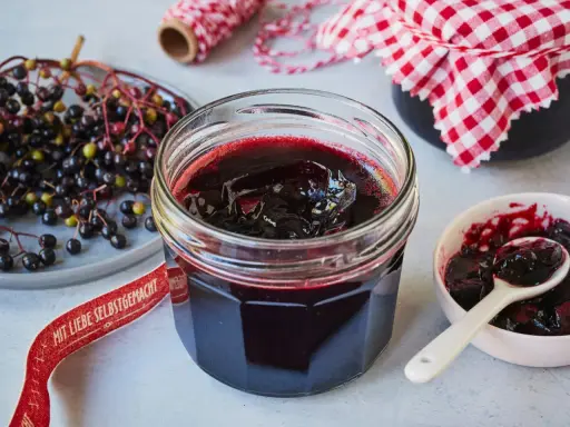 Frisch zubereitete holunderbeeren marmelade im Glas, daneben frische Beeren und eine kleine Schale mit Löffel.