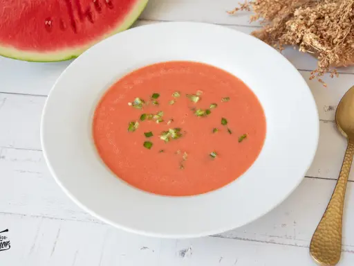 Un refrescante gazpacho de sandía, adornado con trocitos verdes, listo para disfrutar junto a una rodaja de sandía y una cuchara dorada.