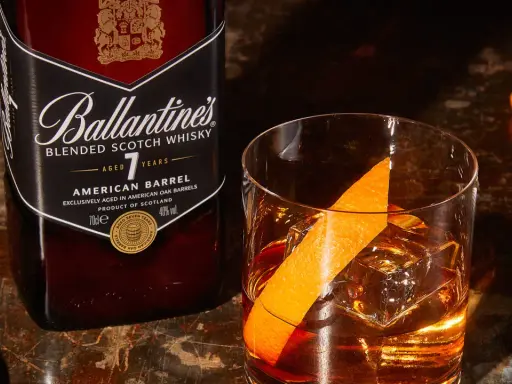 Orzeźwiający drink: whisky z wodą gazowaną, lodem i skórką pomarańczy, obok butelka Ballantine's.