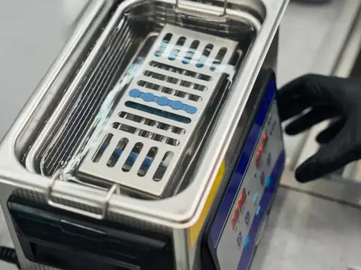 Ein Handschuh bedient ein Gerät, das zeigt, wie funktioniert ein ultraschallreiniger. Kleine Instrumente liegen in einem Korb im Wasserbad.