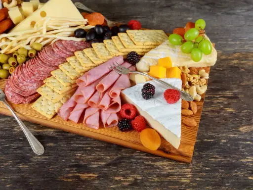 Apetyczna deska serów i wędlin: salami, szynka, ser żółty, pleśniowy, oliwki, winogrona, maliny, orzechy i krakersy.