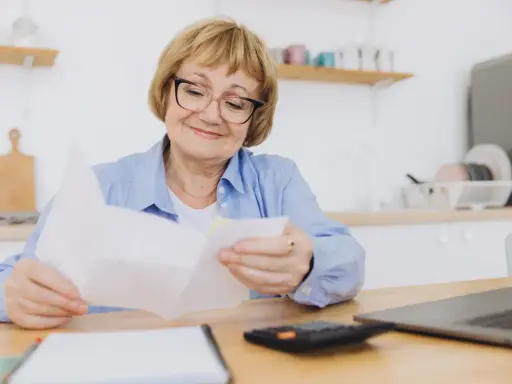 Uśmiechnięta starsza pani z okularami przegląda dokumenty, planując swoje przejście na emeryturę. Obok leży kalkulator i laptop.