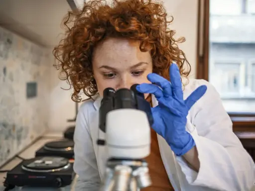 Badania do hematologa: młoda kobieta w niebieskich rękawiczkach bada próbkę pod mikroskopem.