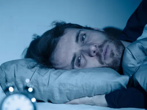 Mann liegt wach im Bett, Augen weit offen, die Angst vor Schlaflosigkeit ist ihm ins Gesicht geschrieben. Ein Wecker ist im Vordergrund zu sehen.