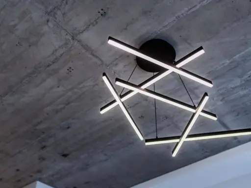Oryginalna lampa sufitowa z geometrycznymi, podświetlanymi ramionami, zawieszona na tle surowego, betonowego stropu.