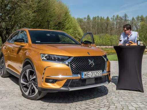 Pomarańczowy Citroën DS7 Crossback z otwartymi drzwiami, obok stolika z winem i bagietką, na tle zieleni.