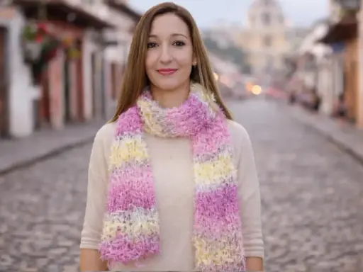 Mujer sonriente luce bufandas de dos agujas modernas en tonos pastel, paseando por una calle empedrada.