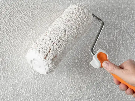 Eine Hand mit orangefarbenem Griff hält eine Farbrolle, die weißen Putz auf eine strukturierte Wand aufträgt. Der Anschluss Putz an Holz wird hier perfektioniert.