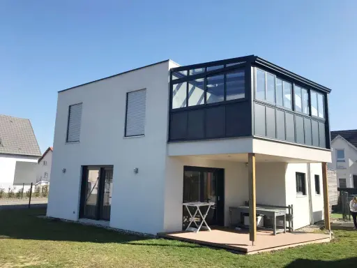 Maison moderne avec extension étage sur piloti. Une véranda vitrée surplombe une terrasse en bois.