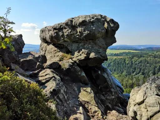 Wielkie, szare skały górują nad zieloną doliną. W oddali widać pola i lasy, a na niebie błękit i chmury.