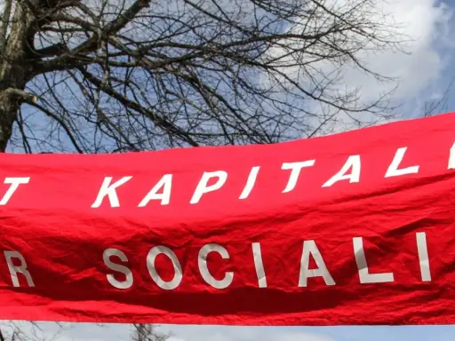 Röd banderoll med vit text: "MOT KAPITALIS FÖR SOCIALIS". En demonstration som visar vad socialism betyder.