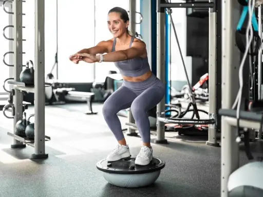 Frau macht Kniebeugen auf einem Bosu-Ball, trägt Sportkleidung und weiße Sneaker. Sie trainiert ohne schuhe auf dem instabilen Untergrund.