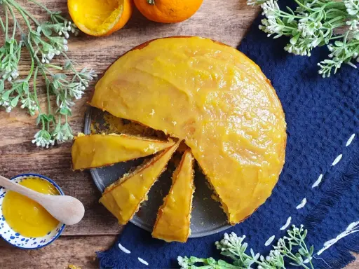 Una torta all'arancia morbidissima, con glassa lucida e profumata, pronta per essere gustata. Accanto, una ciotolina con crema all'arancia e un cucchiaino di legno.