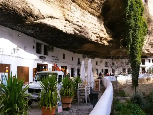 Häuser unter Felsvorsprung in Setenil de las Bodegas. Ein Auto und Pflanzen vor dem "El Gastor" Bar.
