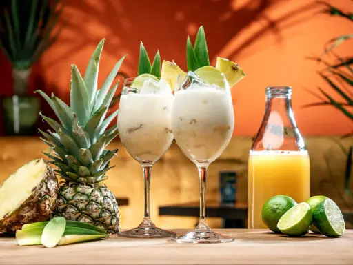 Två krämiga drinkar med ananasjuice, is och lime. En hel och en halv ananas samt en flaska juice står bredvid.