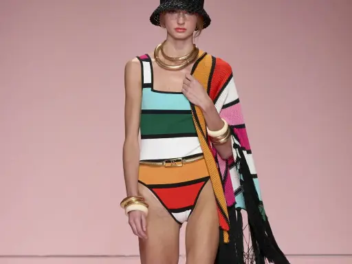 Modella con costume a righe colorate e cappello nero. Un'abbronzatura a strisce perfetta per l'estate, completata da gioielli dorati e un coprispalle a fantasia.