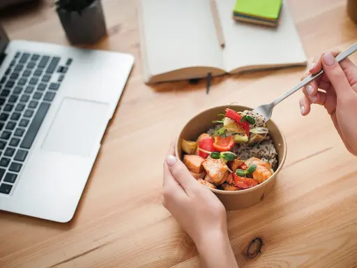 Ręce trzymające miskę z łososiem i warzywami, obok laptop i notatnik. Idealny posiłek na dieta okienkowa.