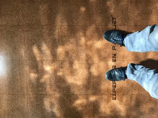 Pintura para corcho con un diseño de baldosas y sombras de luz. Se ven unos pies con zapatos oscuros y pantalones vaqueros.