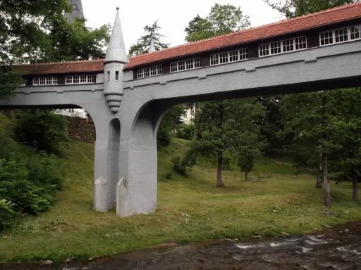 Mostek z wieżyczką, jedna z atrakcji Lądka Zdroju, przerzucony nad strumieniem, otoczony zielenią.
