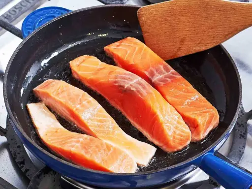 Vier Lachsfilets braten in einer Pfanne. Ein Holzspatel liegt bereit, um zu zeigen, wie bereite ich Lachs zu.