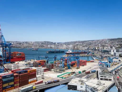 Un vasto porto con gru, container colorati e navi cargo, con una città collinare sullo sfondo. Sembra il porto più grande del mondo.