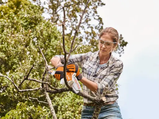 Femme en lunettes de protection et gants orange, effectuant l'entretien des arbres avec une tronçonneuse.