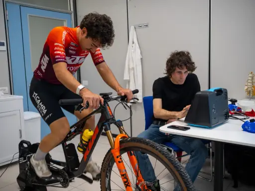 Ciclista su rulli monitorato per l'acido lattico, con un tecnico che osserva i dati su un dispositivo.