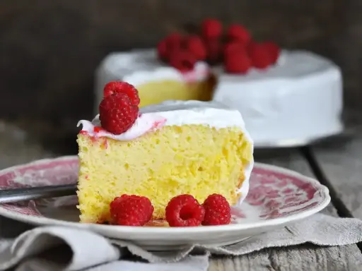 Wilgotne ciasto tres leches z malinami na talerzyku, w tle cały tort.