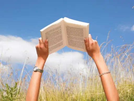 Mani che sorreggono un libro aperto contro il cielo azzurro, tra fili d'erba alta. Un momento di pace e apprendimento, magari con libri sulla salute mentale.