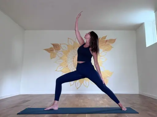Kvinna i krigarställning i ett ljust rum med solrosmålning på väggen. En lugn yoga andrum.