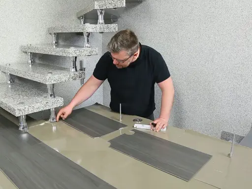 Mann verlegt PVC-Boden. Die grauen Planken werden passgenau zugeschnitten und neben einer modernen Treppe ausgelegt.