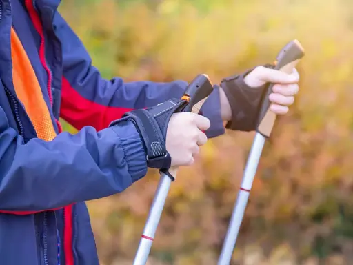 Osoba w kurtce ćwiczy, jak chodzić z kijkami trekkingowymi. Dłonie pewnie trzymają uchwyty, gotowe do marszu.