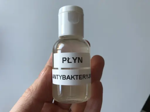 Dłoń trzyma małą, przezroczystą butelkę z białym zamknięciem, zawierającą płyn.