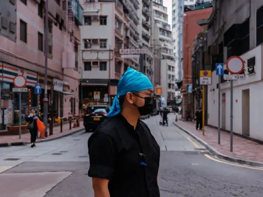 Uomo con bandana blu e mascherina nera in una foto di strada con edifici alti e traffico.