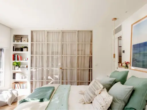 Estantería blanca aprovecha hueco entre cama y pared, con libros y plantas. Puertas de cristal con cortinas separan el espacio.