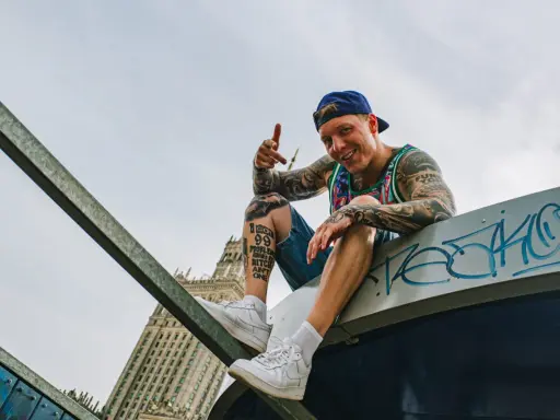 Uśmiechnięty raper z tatuażami, w czapce i kolorowej koszulce, siedzi na dachu, promując swój mixtape "sos ciuchy i borciuchy".